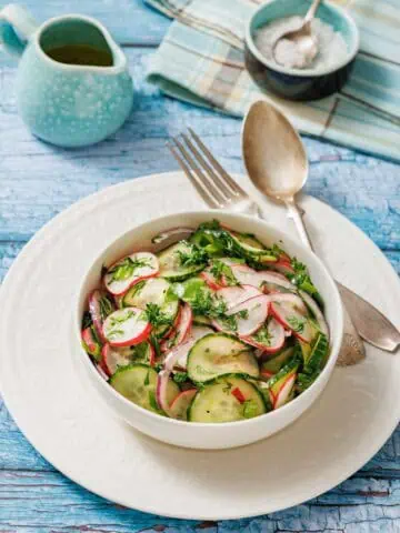Bowl of Radish Salad with herbs on a white plate. Vintage fork and spoon on the side. Light blue wooden table background with a small jug and a napkin.