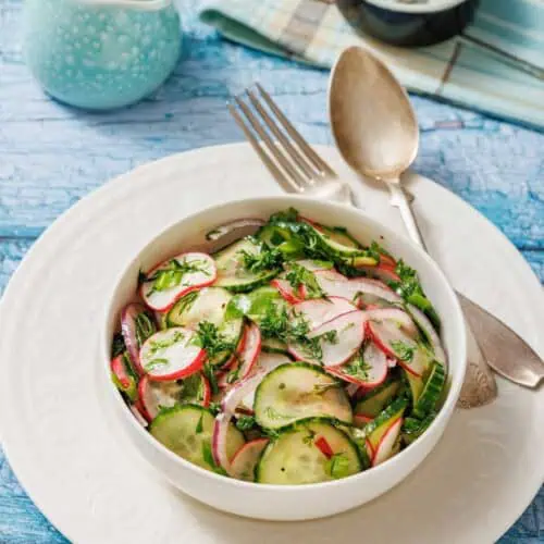 Bowl of Radish Salad with herbs on a white plate. Vintage fork and spoon on the side. Light blue wooden table background with a small jug and a napkin.