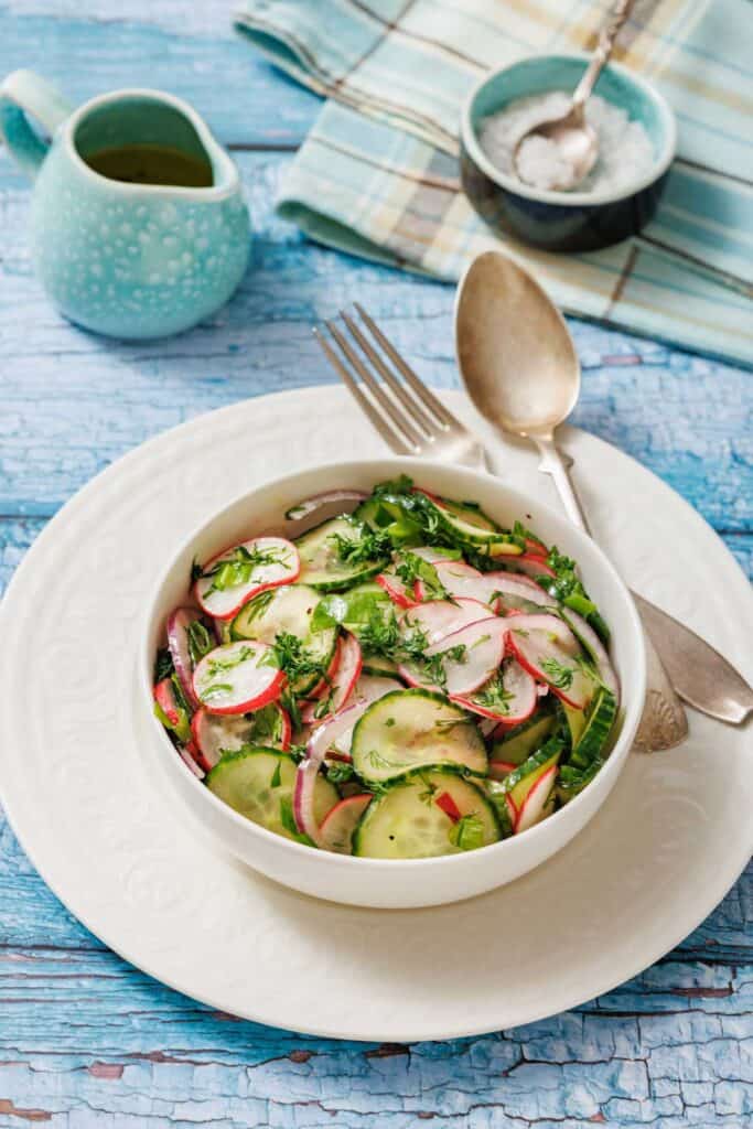 Bowl of Radish Salad with herbs on a white plate. Vintage fork and spoon on the side. Light blue wooden table background with a small jug and a napkin.