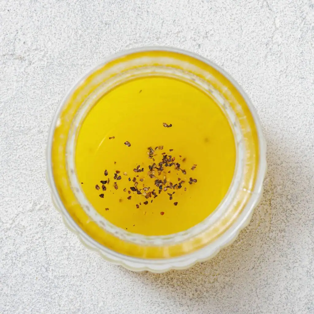 Top view of a transparent bowl filled with yellow liquid, possibly oil, topped with a sprinkling of black pepper on a light textured surface.