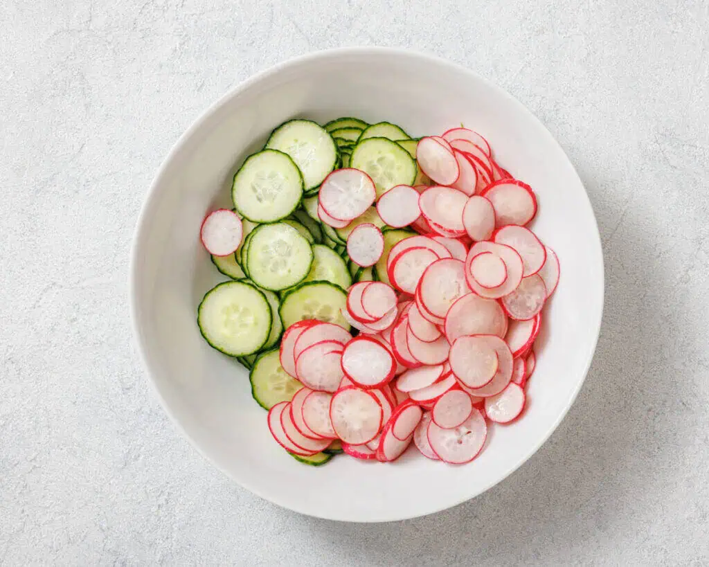 A white bowl with sliced cucumbers and radishes placed side by side on a light gray background.