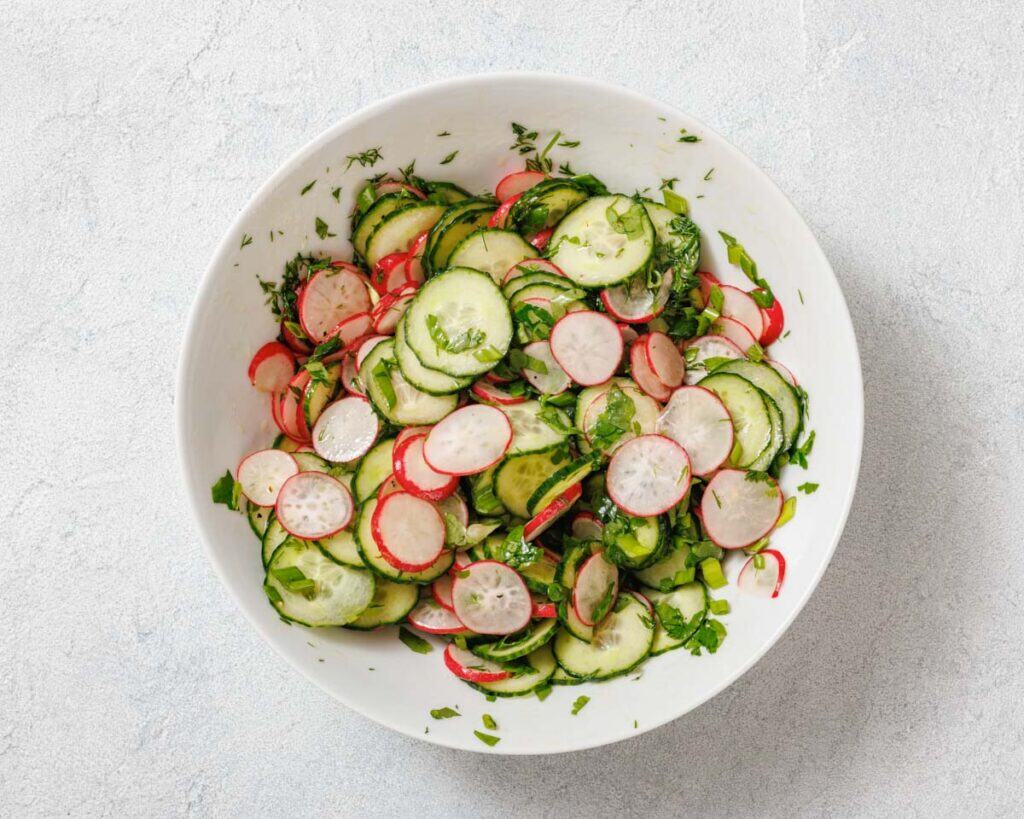 A bowl of fresh salad with sliced cucumbers, radishes, and herbs on a light surface.