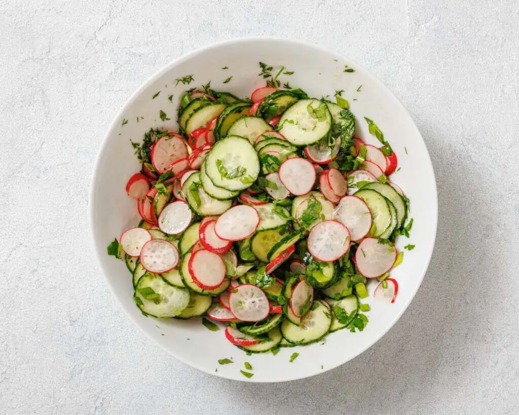 A bowl of fresh salad with sliced cucumbers, radishes, and herbs on a light surface.