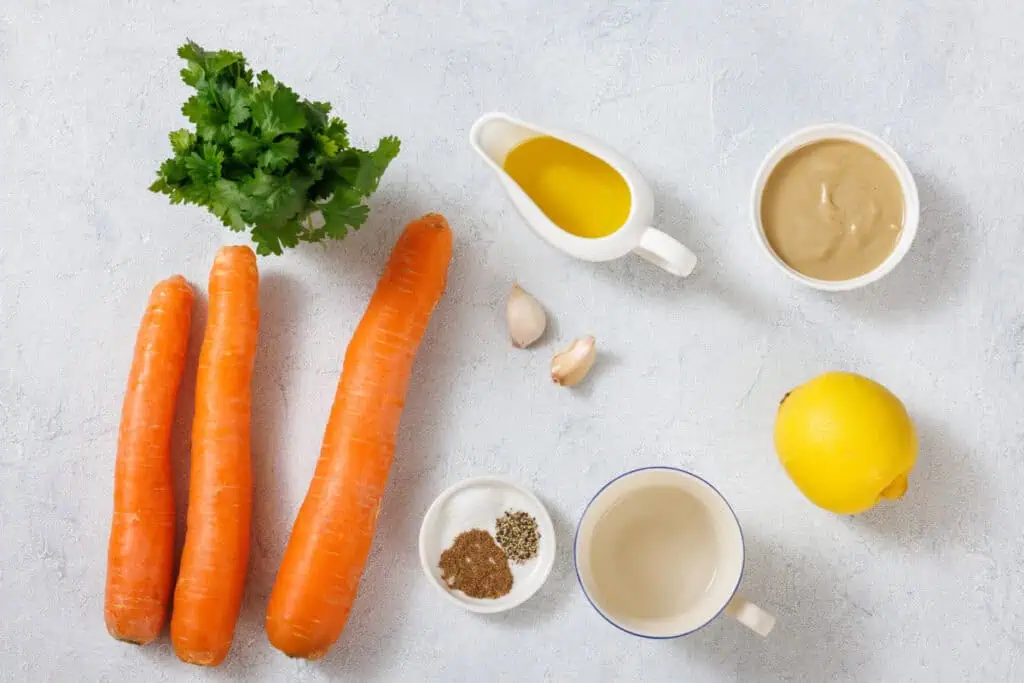 Carrots, cilantro, olive oil, tahini, garlic, lemon, water, and spices are arranged on a light surface.