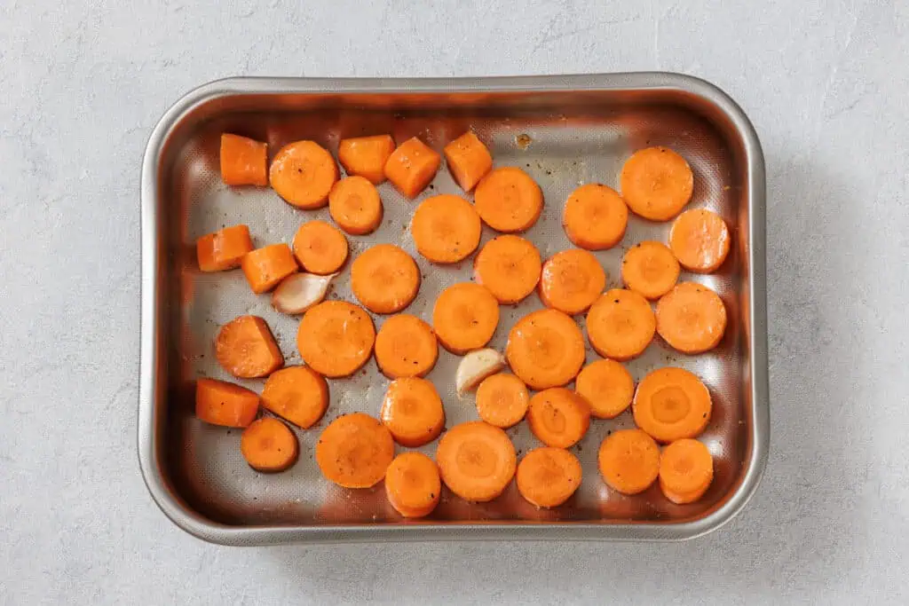 Sliced carrots and garlic cloves seasoned and placed in a metal baking tray on a light gray surface.