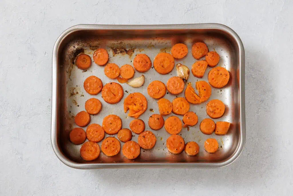 A metal baking tray with sliced roasted carrots and garlic cloves, seasoned with herbs, on a light gray surface.