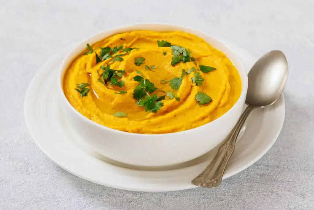 A white bowl of Roasted Carrot Hummus garnished with fresh cilantro, placed on a white plate with a spoon beside it.