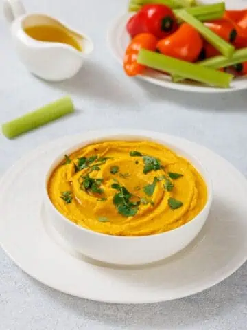 A bowl of Roasted Carrot Hummus garnished with cilantro on a white plate. A small jug of olive oil, celery stick, and a plate of cherry tomatoes and mini peppers are in the background.