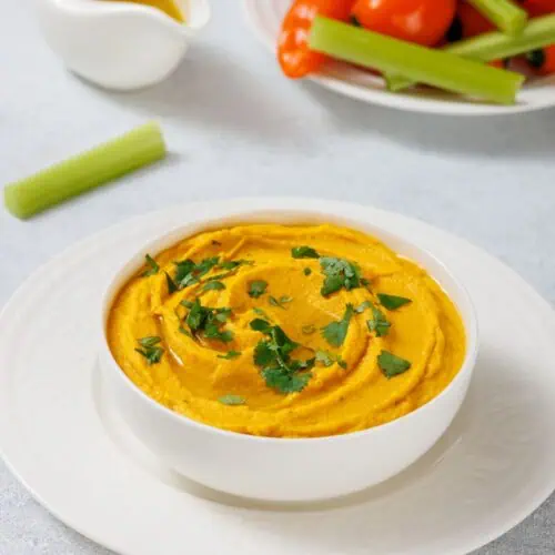 A bowl of Roasted Carrot Hummus garnished with cilantro on a white plate. A small jug of olive oil, celery stick, and a plate of cherry tomatoes and mini peppers are in the background.