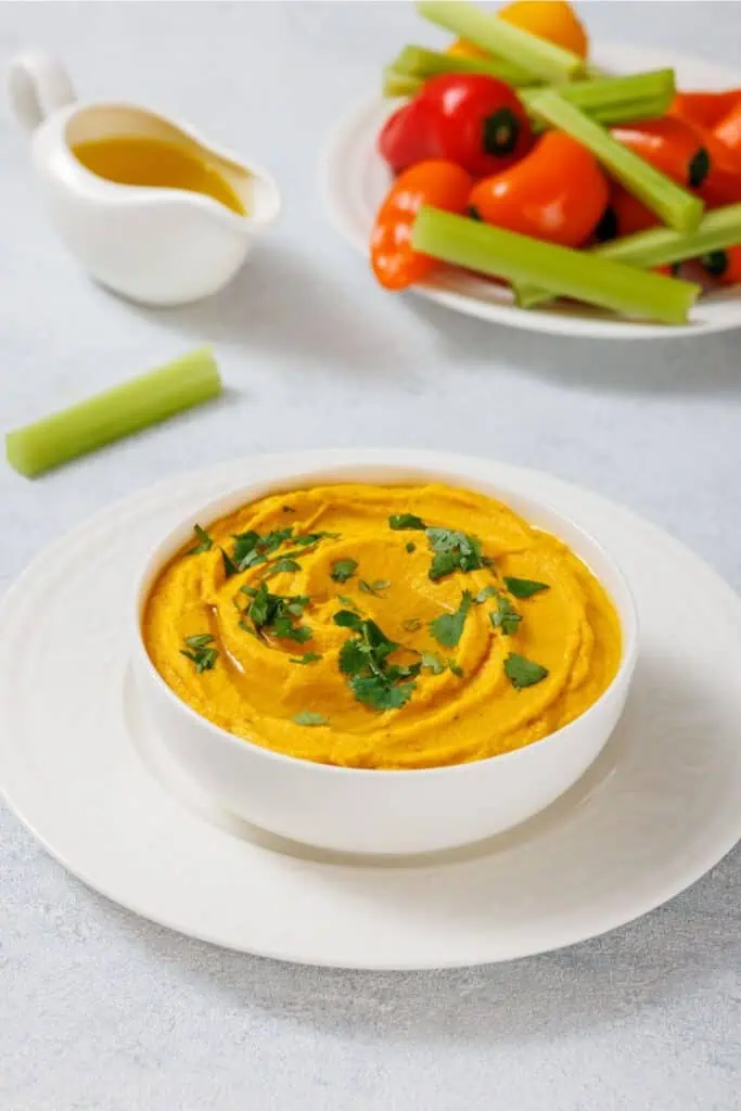 A bowl of Roasted Carrot Hummus garnished with cilantro on a white plate. A small jug of olive oil, celery stick, and a plate of cherry tomatoes and mini peppers are in the background.