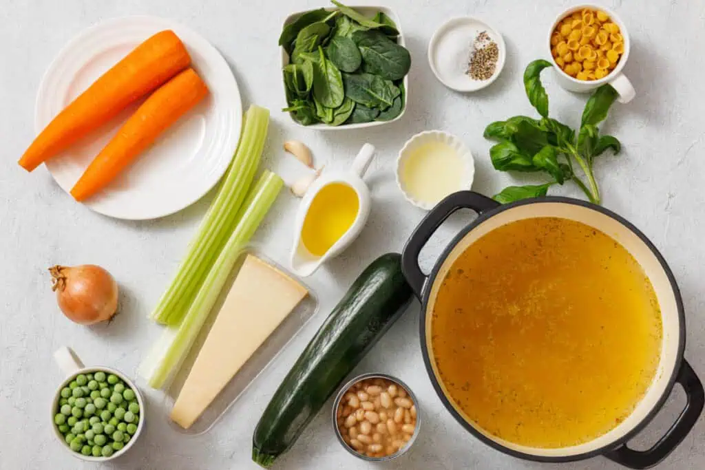 Ingredients for vegetable soup include carrots, spinach, pasta, spices, celery, onion, olive oil, zucchini, peas, cheese, beans, and broth in a pot on a white surface.