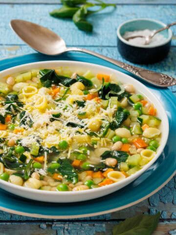 A bowl of Spring Minestrone with pasta, peas, carrots, and spinach, topped with grated cheese. A spoon and salt dish are in the background on a rustic blue surface.