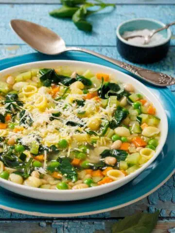 A bowl of Spring Minestrone with pasta, peas, carrots, and spinach, topped with grated cheese. A spoon and salt dish are in the background on a rustic blue surface.