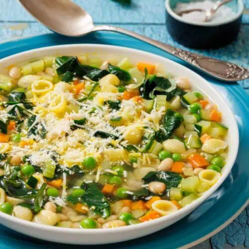 A bowl of Spring Minestrone with pasta, peas, carrots, and spinach, topped with grated cheese. A spoon and salt dish are in the background on a rustic blue surface.