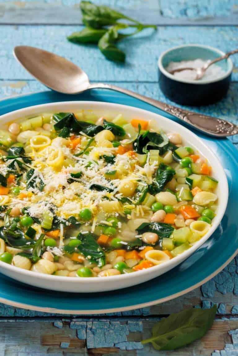 A bowl of Spring Minestrone with pasta, peas, carrots, and spinach, topped with grated cheese. A spoon and salt dish are in the background on a rustic blue surface.