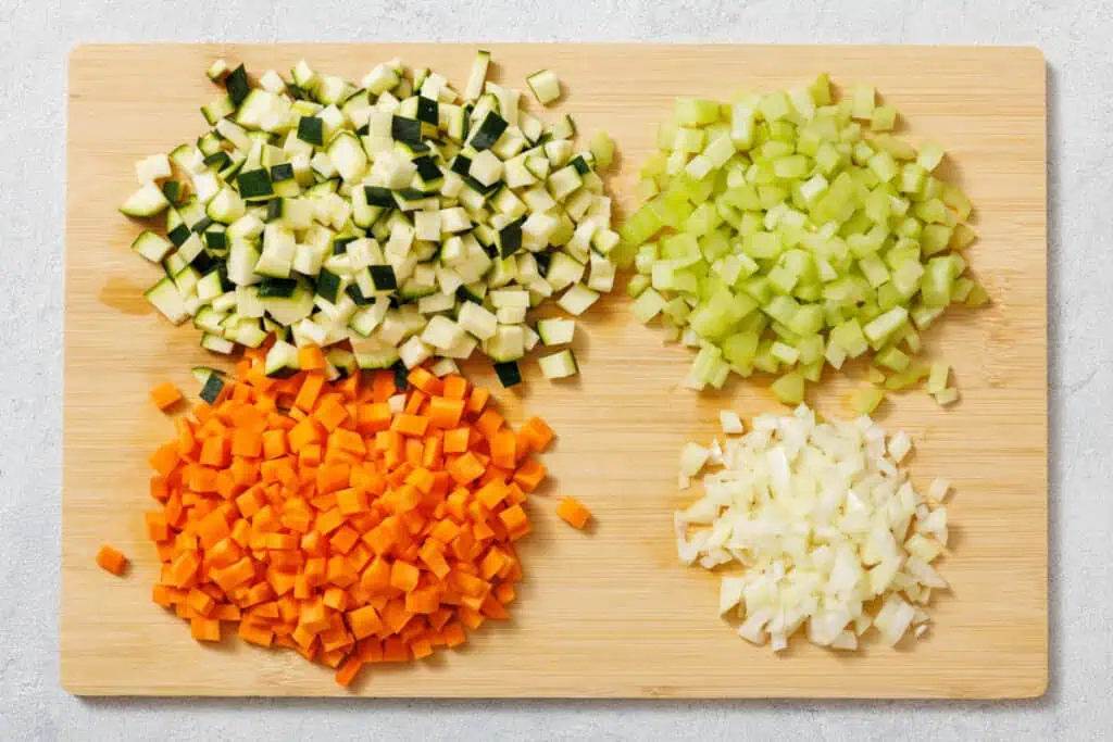 Chopped vegetables on a wooden board: diced zucchini, celery, carrots, and onions, arranged in separate piles.