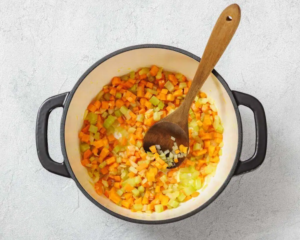 Diced carrots, celery, and onions are being sautéed in a large pot with a wooden spoon on a light surface.