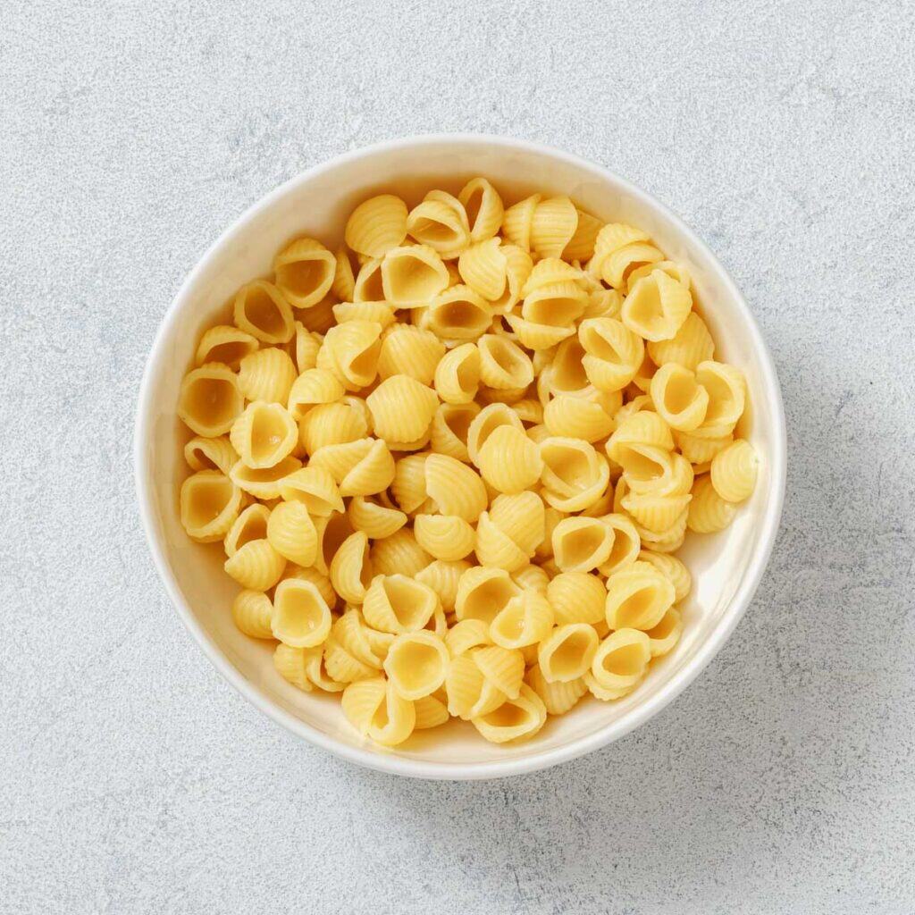 A white bowl filled with uncooked shell-shaped pasta on a light textured surface.