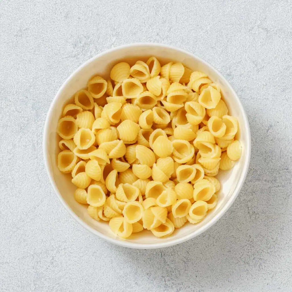 A white bowl filled with uncooked shell-shaped pasta on a light textured surface.
