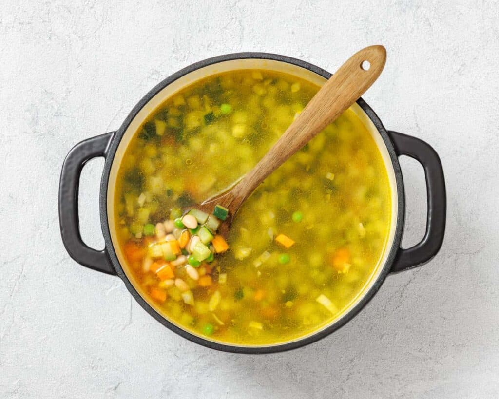 Pot of vegetable soup with a wooden spoon, containing peas, carrots, corn, and green onions, on a textured white surface.