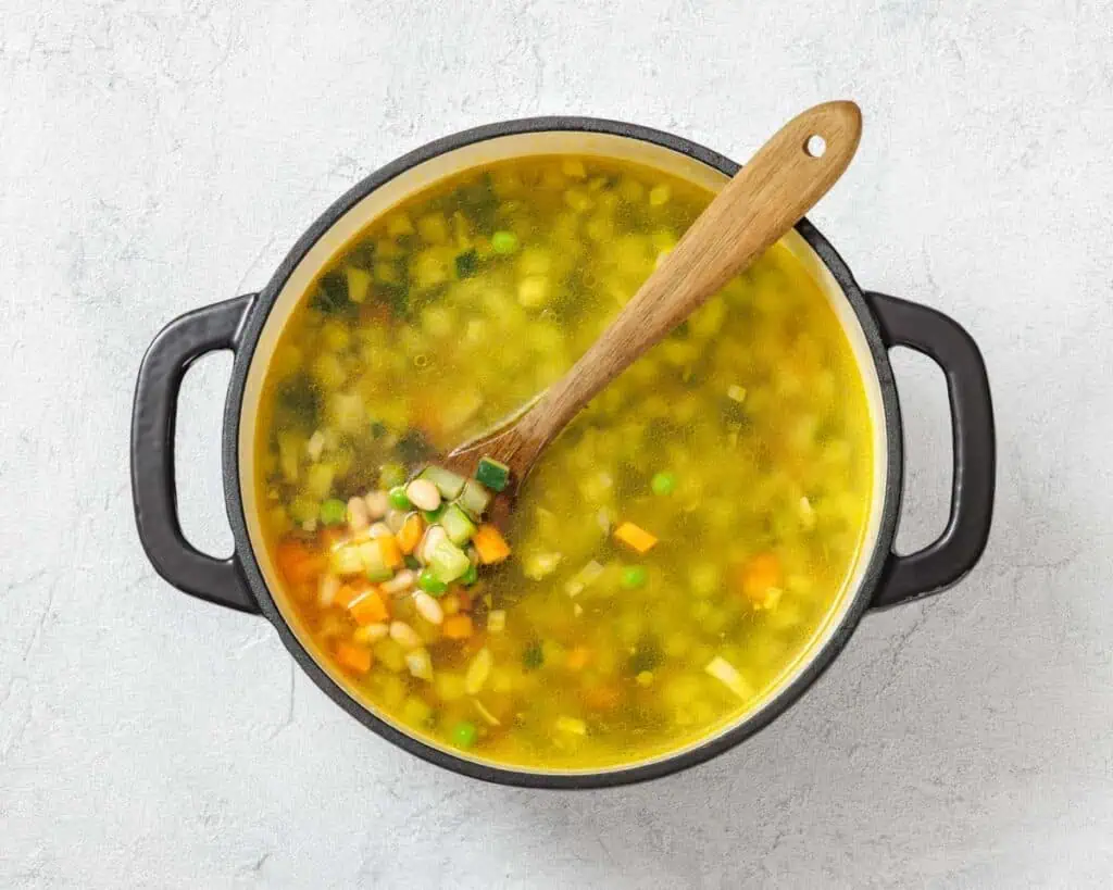 Pot of vegetable soup with a wooden spoon, containing peas, carrots, corn, and green onions, on a textured white surface.