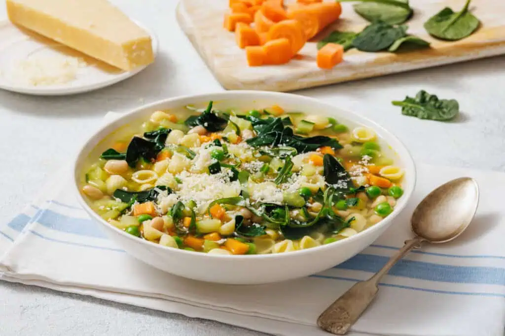 Bowl of Spring Minestrone with pasta, spinach, and beans, topped with grated cheese. A spoon is beside the bowl, and sliced vegetables are on a cutting board in the background.