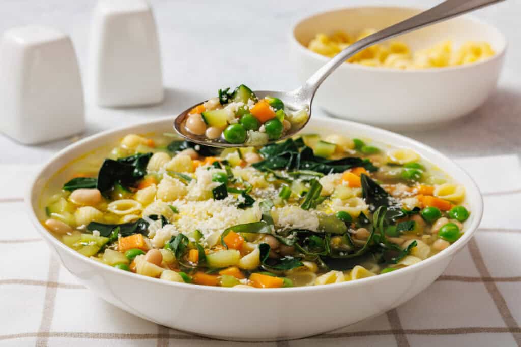 A bowl of Spring Minestrone with pasta shells, peas, carrots, beans, and greens. A spoon hovers above, holding a serving. Salt and pepper shakers in the background.