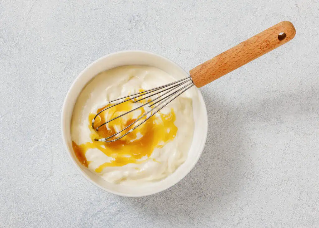 A mixing bowl with yogurt and honey, stirred using a whisk with a wooden handle, placed on a light textured surface.