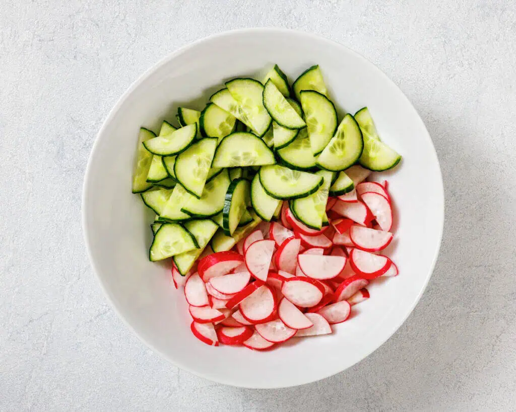 A white bowl filled with sliced cucumbers and radishes on a light gray surface.