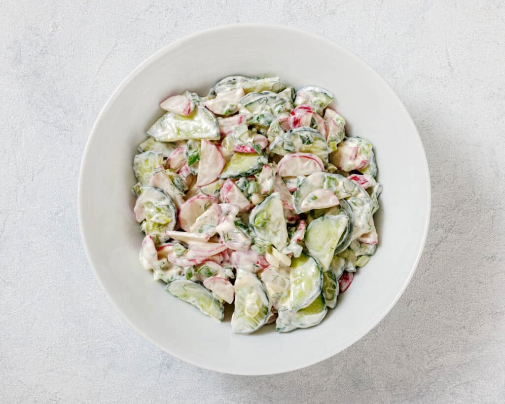 A bowl of creamy cucumber salad with sliced cucumbers, radishes, and herbs on a light background.