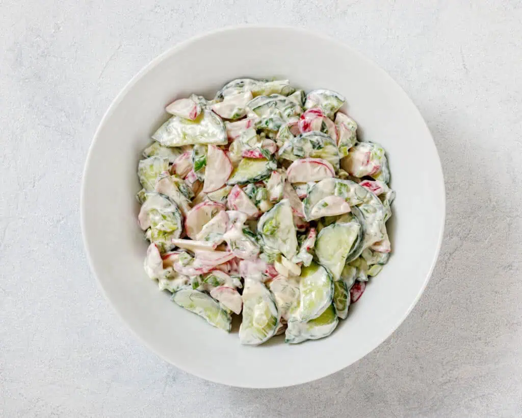 A bowl of creamy cucumber salad with sliced cucumbers, radishes, and herbs on a light background.