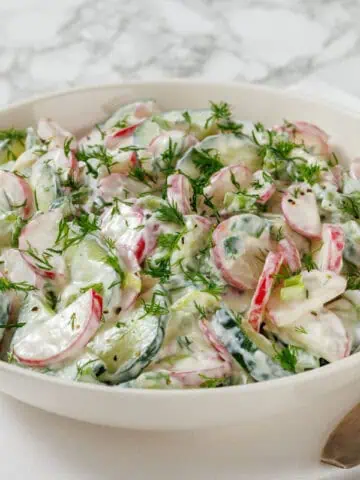 A bowl of Creamy Radish Sala topped with fresh dill, was placed on a white and blue striped napkin with a fork beside it.