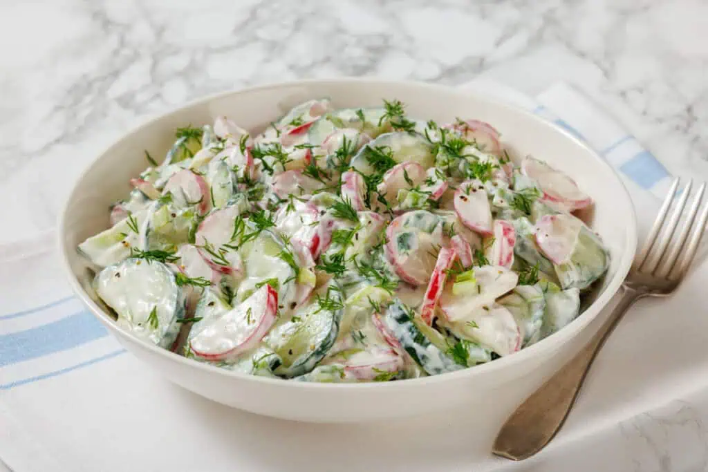 A bowl of cucumber and radish salad with dill and creamy dressing, placed on a white and blue napkin with a fork on the side.