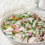 A bowl of fresh salad with sliced radishes, cucumbers, and herbs, dressed in a creamy white sauce, on a white plate.