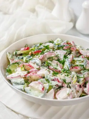 A bowl of fresh salad with sliced radishes, cucumbers, and herbs, dressed in a creamy white sauce, on a white plate.