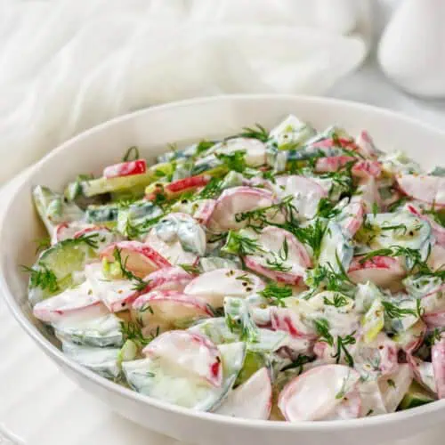 A bowl of fresh salad with sliced radishes, cucumbers, and herbs, dressed in a creamy white sauce, on a white plate.