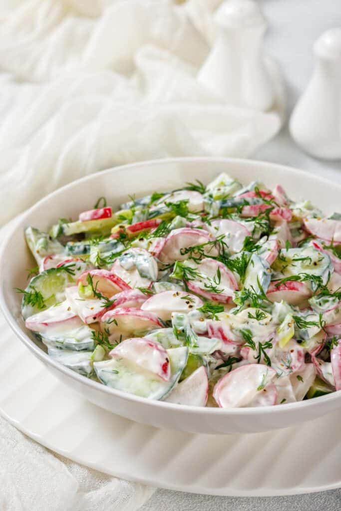 A bowl of fresh salad with sliced radishes, cucumbers, and herbs, dressed in a creamy white sauce, on a white plate.
