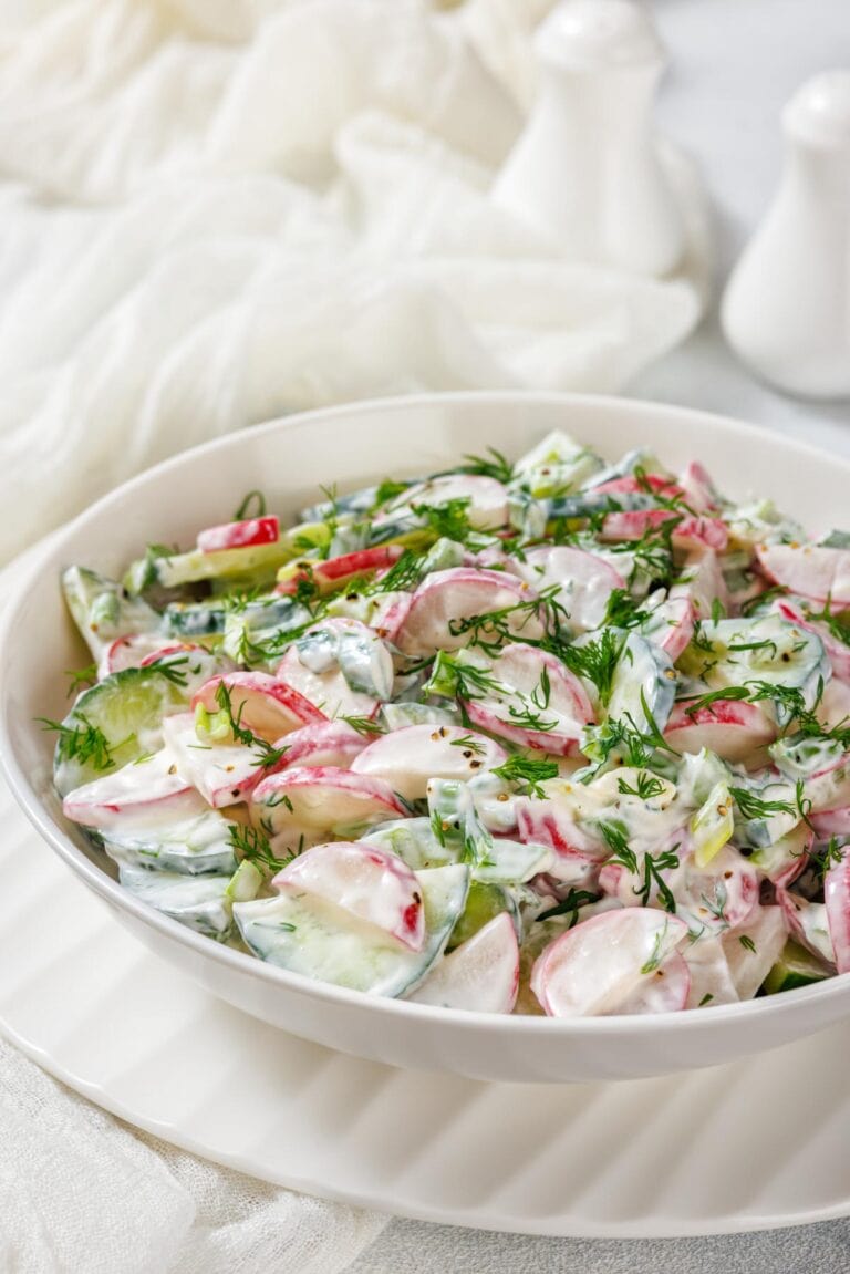 A bowl of fresh salad with sliced radishes, cucumbers, and herbs, dressed in a creamy white sauce, on a white plate.