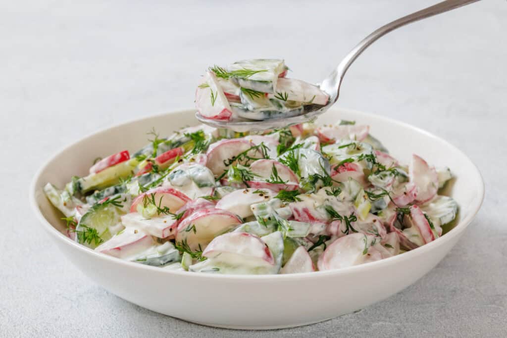 A bowl of creamy salad with sliced radishes, cucumbers, and dill. A spoon holds a portion above the bowl.