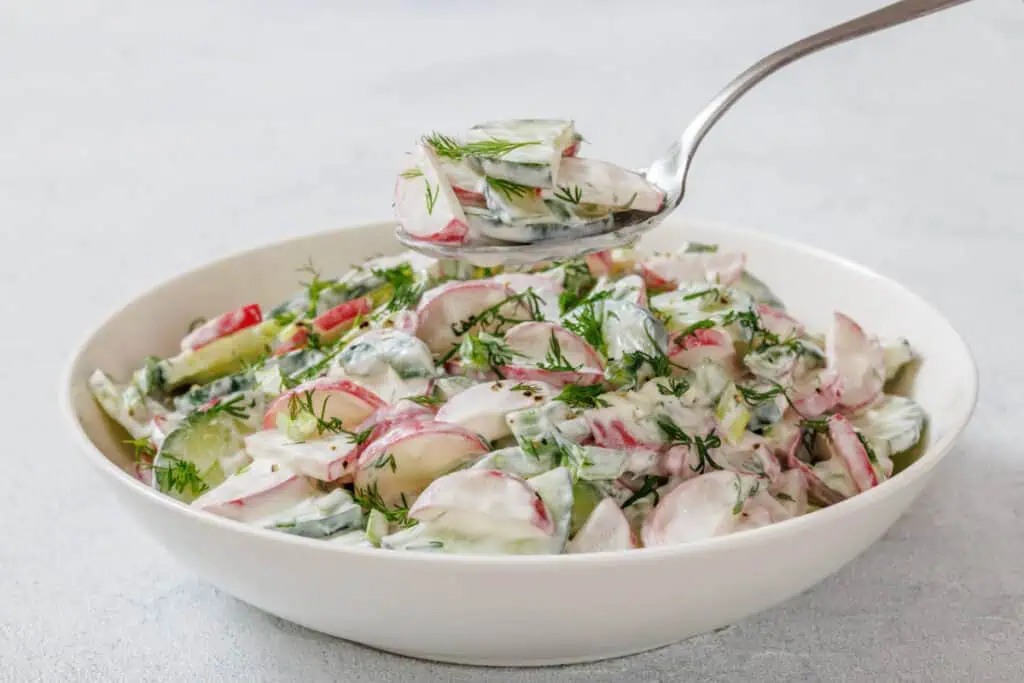 A bowl of creamy salad with sliced radishes, cucumbers, and dill. A spoon holds a portion above the bowl.