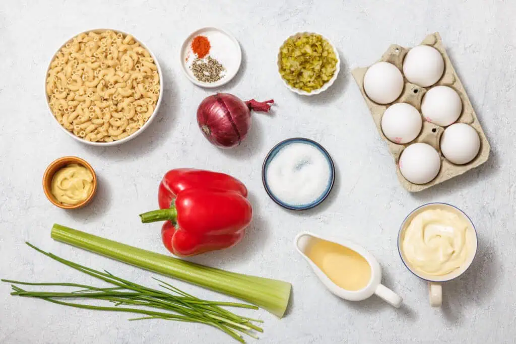 Ingredients for Deviled Egg Pasta Salad arranged on a surface include macaroni, eggs, mayonnaise, red bell pepper, celery, chives, onion, pickles, mustard, and seasonings.