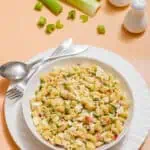 A bowl of Deviled Egg Pasta Salad with vegetables on a white plate, accompanied by a fork and spoon. Background features celery stalks and salt and pepper shakers.