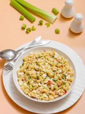 A bowl of Deviled Egg Pasta Salad with vegetables on a white plate, accompanied by a fork and spoon. Background features celery stalks and salt and pepper shakers.