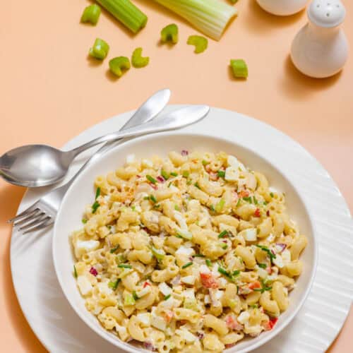 A bowl of Deviled Egg Pasta Salad with vegetables on a white plate, accompanied by a fork and spoon. Background features celery stalks and salt and pepper shakers.