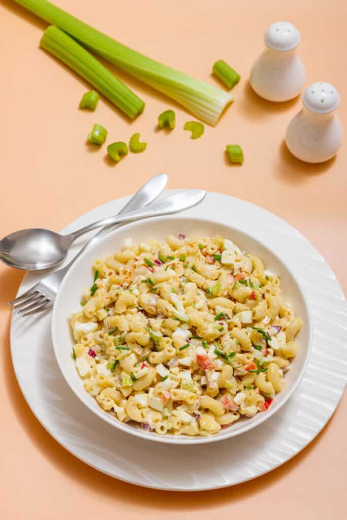 A bowl of Deviled Egg Pasta Salad with vegetables on a white plate, accompanied by a fork and spoon. Background features celery stalks and salt and pepper shakers.