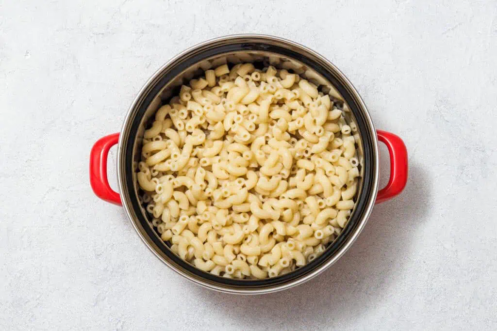 A pot with red handles filled with cooked elbow macaroni pasta on a light textured surface.