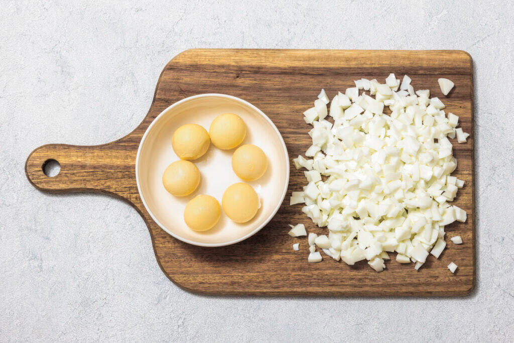 Chopped white onions and six yellow onions in a bowl sit on a wooden cutting board.