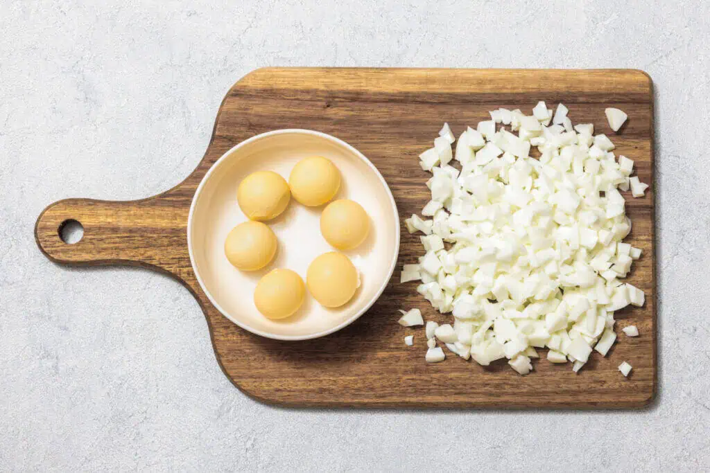 Chopped white onions and six yellow onions in a bowl sit on a wooden cutting board.
