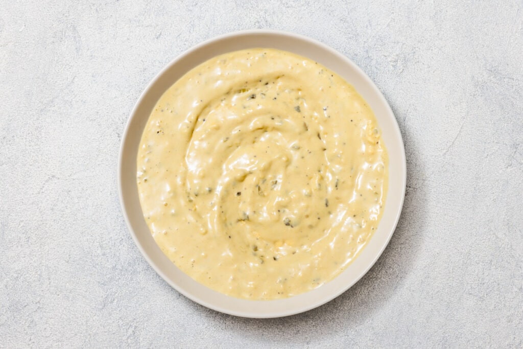 A bowl of creamy, beige sauce with herbs on a light gray textured surface.