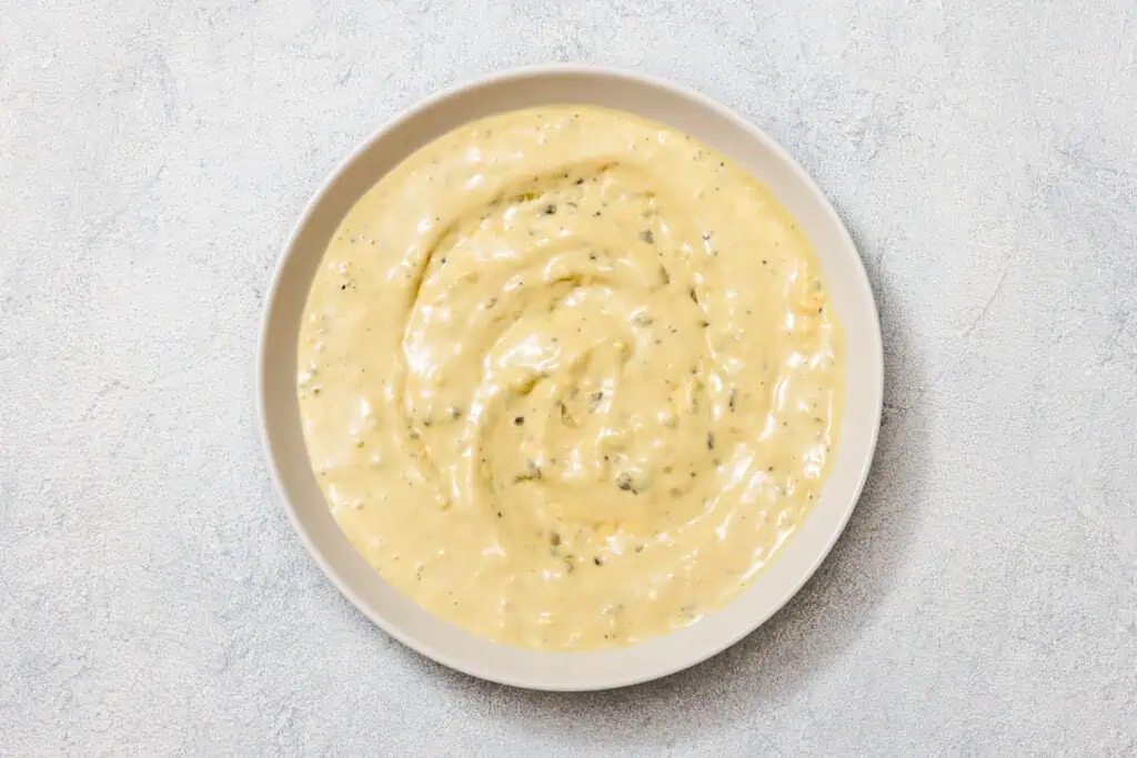 A bowl of creamy, beige sauce with herbs on a light gray textured surface.